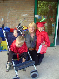 Two twin boys wear red gowns as they graduate nursery. One sits in a push chair.