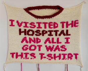 Needlework by Emily Robbins. White square with the neck opening of a t-shirt at the top. Read text reads "I visited the hospital and all I got was this T-Shirt"