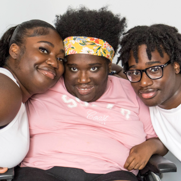 A woman who is a wheelchair user smiles for the camera, with her brother and sister leaning in on each side of her